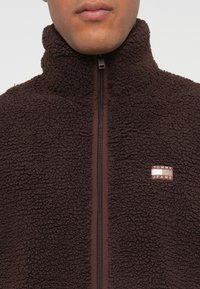 Jachetă din fleece maro, cu guler înalt, fermoar la față și o mică etichetă cu brand pe pieptul stâng. Textură moale și design cald.
