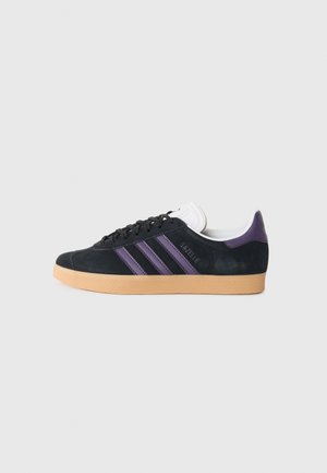 Svarte semskede sneakers med tre lilla striper, en hvit tunge og en gummisåle. Har en rund tå og merke detaljer på hælen.