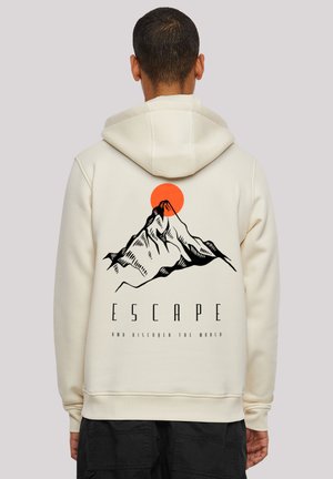 Man wearing cream hoodie with black mountain graphic and red sun above, text "ESCAPE and discover the world" on back.