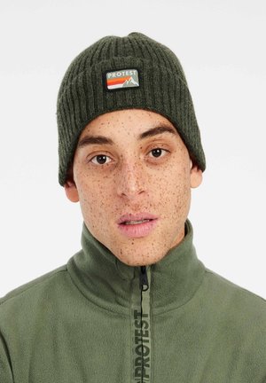 Beanie a coste di colore verde scuro con una patch triangolare che rappresenta una montagna, caratterizzata da dettagli arancioni, bianchi e verdi. Design con corona piatta e rotonda.