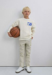 Sudadera color crema con diseño gráfico azul, pantalones cargo beige y zapatillas blancas. Niño sosteniendo un balón de baloncesto, de pie sobre un fondo claro.