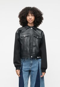 Mustast kunstnahast lühike jakk kahe rinnatasku, klõpsuga sulgemise ja mustade kangaste varrukatega. Koos laia säärega siniste teksadega.