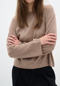 Femme aux longs cheveux bruns, les bras croisés, portant un haut beige texturé à manches longues et un pantalon noir sur un fond clair uni.