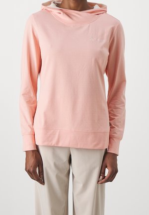 Osoba ubrana w jasnoróżową, długorękawową bluzę z kapturem i beżowe spodnie, stojąca na tle jednolitego białego tła.