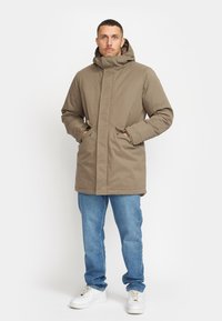 Brun, hættejakke til udendørs brug med lynlåse og trykknap-lukning, med sidelommer og en afslappet pasform, parret med blå jeans og hvide sneakers.