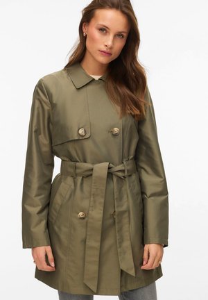 Femme aux longs cheveux châtain portant un trench-coat ceinturé vert olive avec des boutons sur un fond clair.