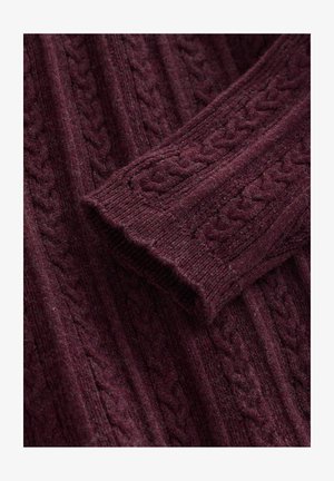 Burgundi színű, kötött gyapjúpulóver ujja a megfelelő pulóver testére hajtva, megjelenítve a texturált kötött mintát és a bordás mandzsettát.