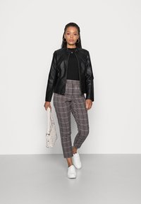 Svart faux skinnjakke, tettsittende svart strikket topp, rutete bukser i brunt og krem, hvite sneakers, vevd beige veske, modell som står på grått gulv.