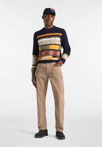 Maglione lavorato a maglia con strisce multicolori e motivo floreale, abbinato a pantaloni in velluto a coste marrone chiaro e stivali neri in camoscio. Berretto blu in testa.