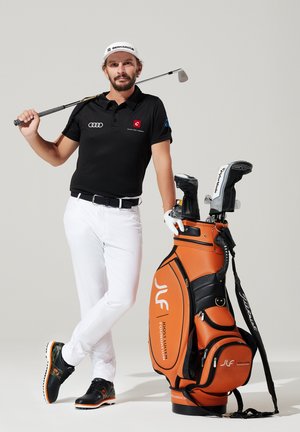 Schwarzes Poloshirt mit Logo, weiße Hose, schwarz-orange Golfschuhe. Oranger Golfbag mit schwarzen Akzenten und Branding. Golfschläger auf der Schulter.