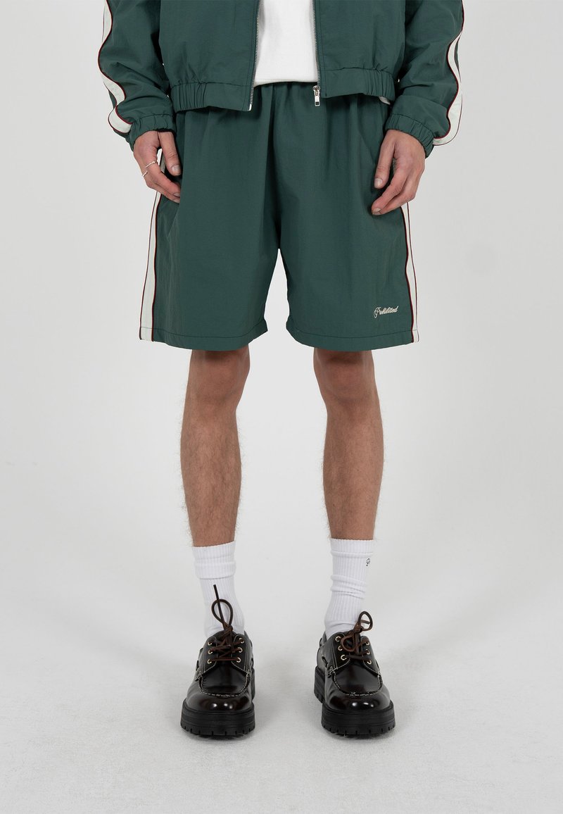 Homme portant un short vert foncé et une veste assortie, des chaussettes blanches et des chaussures en cuir noires, debout devant un fond gris clair.