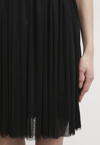 Main droite reposant à côté d'une jupe noire plissée jusqu'au genou avec une superposition de tissu transparent texturé.