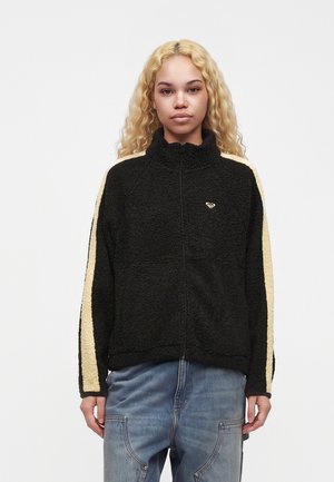Jeune femme aux longs cheveux blonds bouclés porte une veste polaire noire avec des bandes beiges sur les manches et un jean bleu, face vers l'avant.