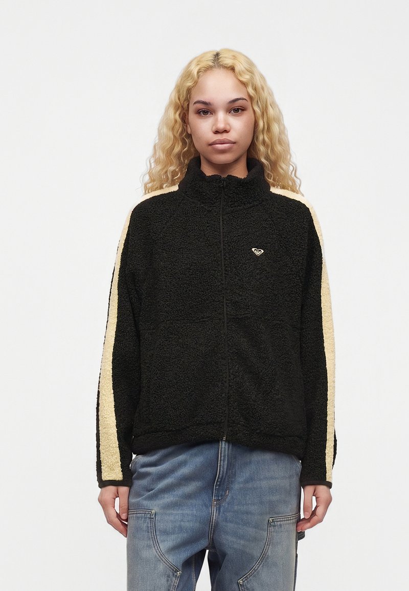 Jeune femme aux longs cheveux blonds bouclés porte une veste polaire noire avec des bandes beiges sur les manches et un jean bleu, face vers l'avant.