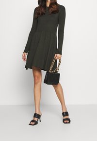 Robe verte côtelée avec des manches longues et un ourlet évasé, associée à un sac à main matelassé noir avec une bandoulière en chaîne dorée et des sandales à talons noires.