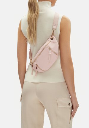 Frau trägt ärmelloses weißes Oberteil und beige Cargo-Hose mit einer hellrosa Umhängetasche, die mehrere goldene Reißverschlüsse hat.