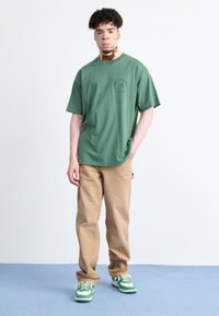 Groen katoenen T-shirt met korte mouwen en een ronde halslijn, voorzien van een kleine grafische print. Gecombineerd met beige broek en groene sneakers.