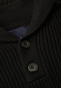 Schwarzer Strickpullover mit gerippter Textur, ausgestattet mit einem Schalkragen und zwei glänzenden schwarzen Knöpfen. Ein einfarbiger navyblauer Stoff ist darunter sichtbar.