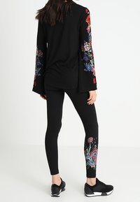 Černý dlouhý rukáv top s květinovou výšivkou na rukávech, kombinovaný s černými legínami s květinovými vzory u kotníků, nošený s černými teniskami.