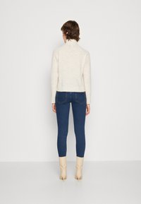 Maglione a maglia color crema con collo alto, abbinato a jeans skinny blu scuro e stivali alla caviglia beige. Tessuto uniforme, silhouette aderente.