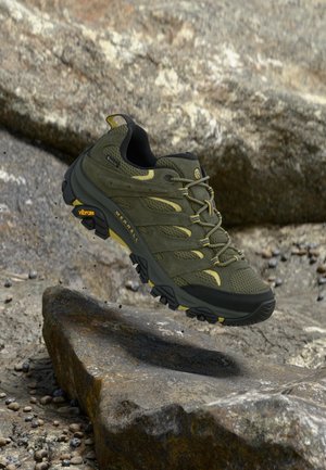 Chaussure de randonnée vert olive et noir avec semelle Vibram, lacets en suspension au-dessus d'un terrain rocheux en plein air.