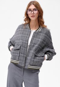 Szara bomberka w kratę z prążkowanymi zielonymi i białymi mankietami, kieszeniami bocznymi i zapięciem na zamek. Stylizowana z białą bluzką i szarymi spodniami.