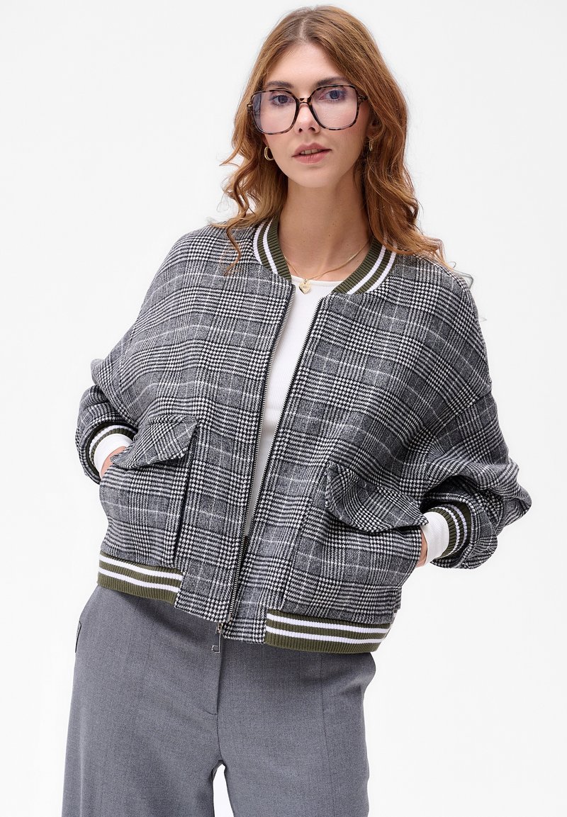 Szara bomberka w kratę z prążkowanymi zielonymi i białymi mankietami, kieszeniami bocznymi i zapięciem na zamek. Stylizowana z białą bluzką i szarymi spodniami.