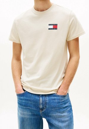 Homme portant un t-shirt crème Tommy Jeans avec un logo sur la poitrine et un jean bleu, les mains dans les poches, debout devant un fond blanc.