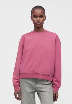 Frau mit kurzen Haaren, die einen schlichten rosa Pullover und hellgraue Jeans trägt, steht vor einem weißen Hintergrund.