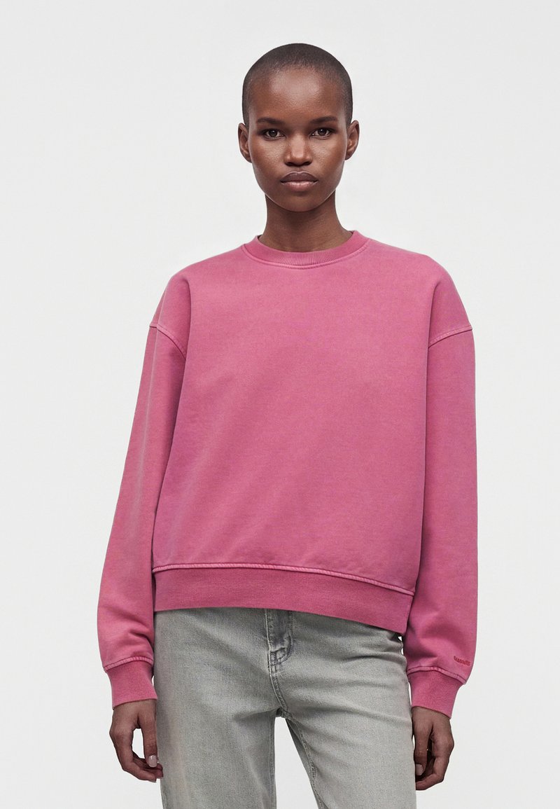 Femme aux cheveux courts portant un sweat-shirt rose uni et un jean gris clair, debout devant un fond blanc.