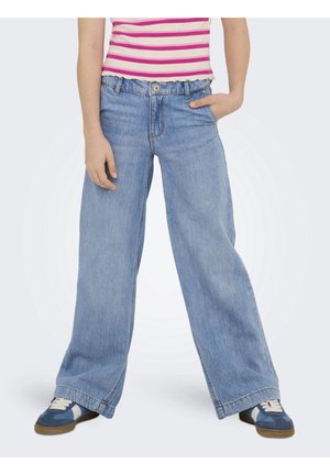 Personne portant un jean bleu clair à jambes larges, des baskets bleu et blanc, et une chemise rose et blanche à rayures avec une main dans la poche.