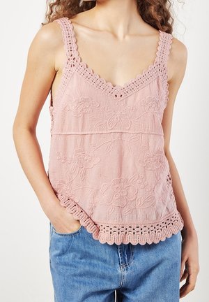 Vrouw draagt een roze mouwloze top met gehaakte rand en bloemenpatronen, gecombineerd met blauwe spijkerbroek, hand in de zak.