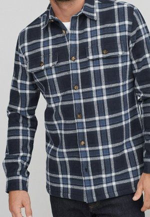 Man draagt een overhemd met lange mouwen, marineblauw en wit geruit, met twee borstzakken en bruine knopen.