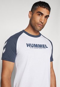 Bílý tričko s námořnickými raglánovými rukávy, se značkovým logem "HUMMEL" ve tmavě modrém písmu. Hladká látka se kontrastním detailem rukávů.