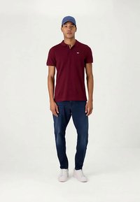 Jovem de pé, vestindo uma camisa polo bordô, jeans azuis, tênis brancos e um boné de beisebol azul, diante de um fundo claro.