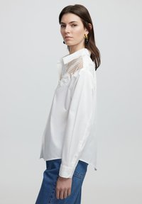 Weißes Langarmshirt mit Spitzen- und Netzdetails an den Schultern, mit Knopfmanschetten und entspannter Passform, kombiniert mit blauen Denim-Hosen.