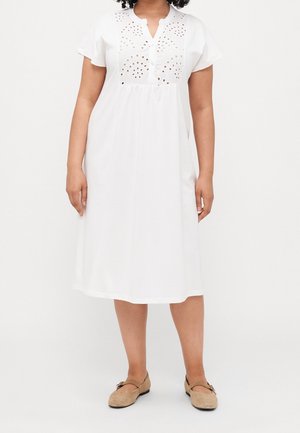 Vrouw die een witte knielange jurk draagt met oogjesuitsparingen op de borst, korte mouwen en beige platte schoenen met gespen.