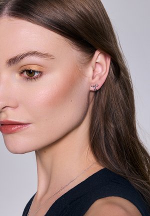 Srebrne kolczyki w kształcie obręczy z motywem kokardy, prezentujące wypolerowane wykończenie. Modelka ma gładką cerę i długie, proste włosy.