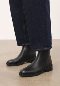 Mustad mustad nahast pahkluu saapad, millel on tumedat sinist sirget jalga teksaseid, sileda beeži põranda peal.