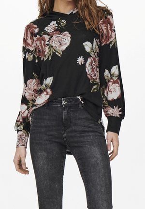 Vrouw met een zwarte blouse met lange mouwen en een roze-witte bloemenprint, ingestopt in een hooggekoekte, vervaagde zwarte spijkerbroek.