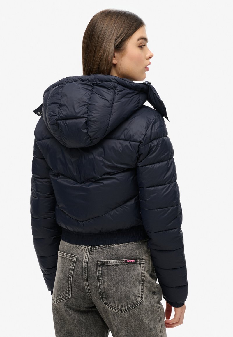 Marineblauwe pufferjack met een capuchon, met een gewatteerde textuur en ribgebreide zoom, gecombineerd met lichtgrijze jeans. Inclusief een subtiel logo-detail op de zak.