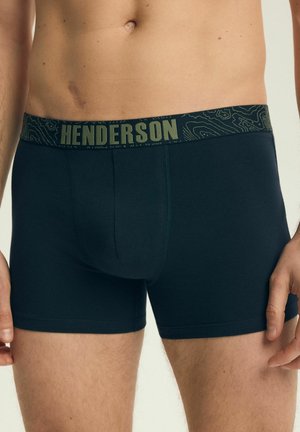 Zbliżenie torsu mężczyzny noszącego czarne bokserki z wzorzystym pasem w talii oznaczonym napisem "HENDERSON".