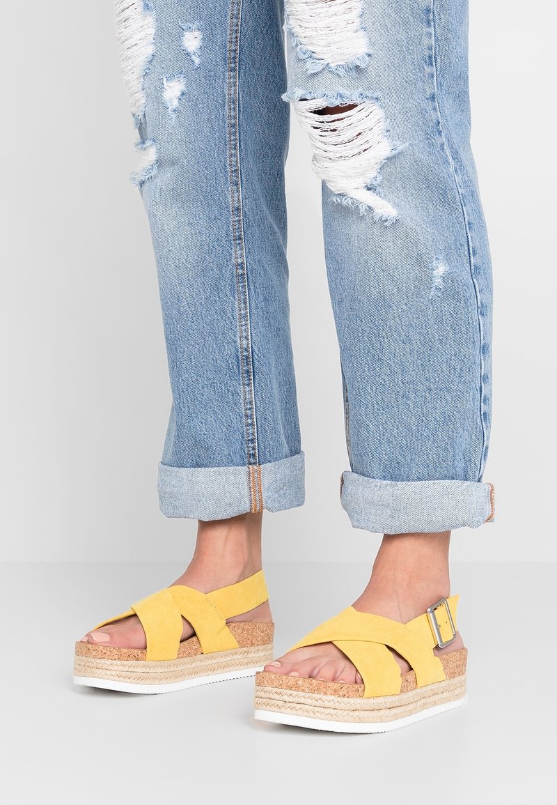 Pieds portant des sandales à plate-forme jaunes avec semelles en liège et lanières à boucles argentées, associées à un jean bleu clair déchiré et retroussé.