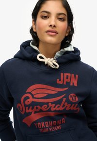 Marineblauwe hoodie van katoenmix, met een rode grafische print met "Superdry" logo, trekkoord hals en gestreepte binnenvoering.
