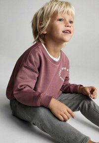 Hanorac cărămiziu cu text alb, cu guler tip crew și manșete elastice, asociat cu pantaloni gri elastici. Copilul este așezat.