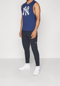 Laineetön tummansininen toppi, jossa on valkoinen logo, yhdistettynä mustiin kapeneviin jogger-housuihin ja valkoisiin tennareihin neutraalilla taustalla.