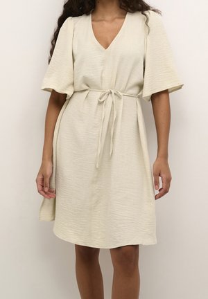 Kobieta ubrana w beżową sukienkę do kolan z krótkimi, szerokimi rękawami, dekoltem w serek i przewiązanym paskiem w talii, stojąca na jednolitym tle.