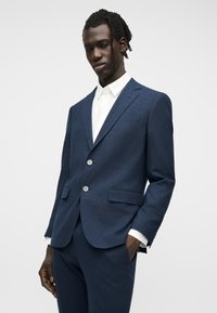 Άνδρας με σκούρο μπλε κοστούμι με δύο κουμπιά και λευκό πουκάμισο, στέκεται με το ένα χέρι στην τσέπη μπροστά σε μονόχρωμο φόντο.
