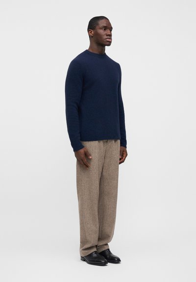 Μπλε πουλόβερ με στρογγυλή λαιμόκοψη, σε συνδυασμό με ανοιχτό καφέ υφασμάτινο παντελόνι και μαύρα παπούτσια, φορεμένα από μια όρθια φιγούρα.