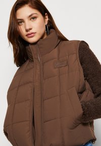 Gilet imbottito marrone trapuntato con colletto alto, zip frontale e tasche laterali; presenta un tessuto marrone testurizzato sulle maniche e una fascia con logo.